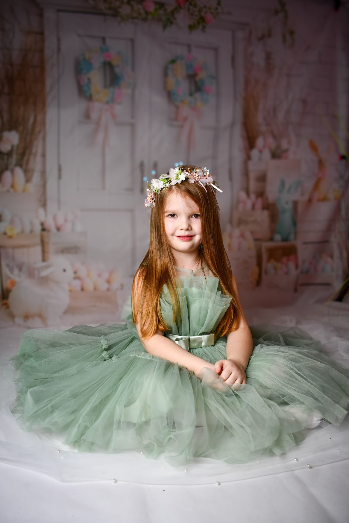 Dziewczynka w tiulowej sukience w kolorze szałwii i wianku z kwiatów na głowie, siedząca na białym tle z dekoracjami wielkanocnymi w tle.