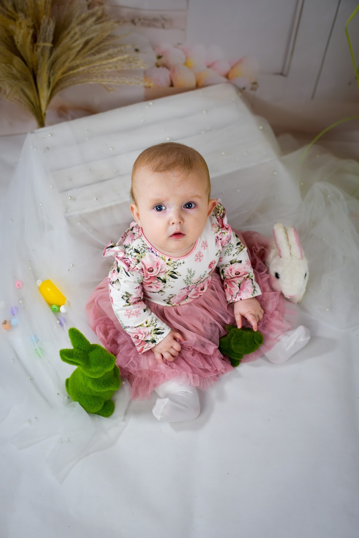 Niemowlę w kwiecistej sukience i tiulowej spódniczce siedzi na białym tle z dekoracjami wielkanocnymi, w tym pluszowym króliczkiem i zielonymi zajączkami z mchu.