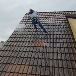 TECH-GARDEN Jacek Kubacki - Pracownik w uprzęży, myjący dach z brązowej dachówki za pomocą myjki ciśnieniowej; widoczny fragment dachu przed i po czyszczeniu.