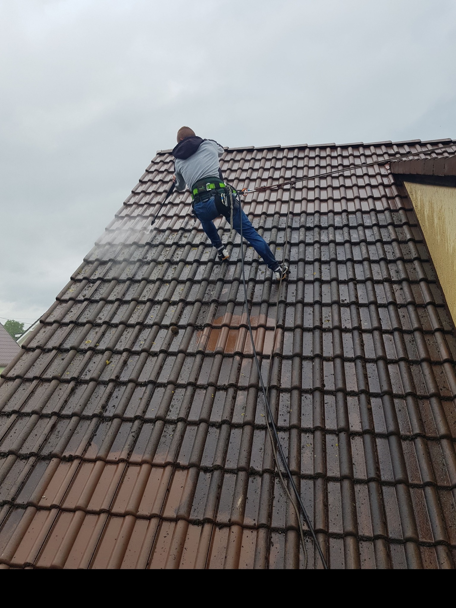 Pracownik w uprzęży, myjący dach z brązowej dachówki za pomocą myjki ciśnieniowej; widoczny fragment dachu przed i po czyszczeniu.