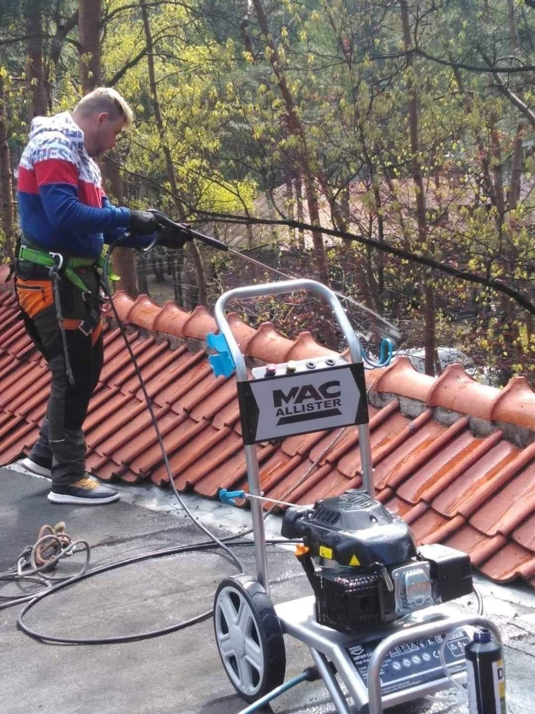 Pracownik w uprzęży bezpieczeństwa czyści dachówkę za pomocą myjki ciśnieniowej Mac Allister, w tle drzewa z młodymi liśćmi.