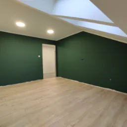 Poddasze po remoncie: ściany pomalowane na ciemnozielony kolor, jasne panele podłogowe, widoczne punkty oświetleniowe w suficie i fragment okna dachowego.