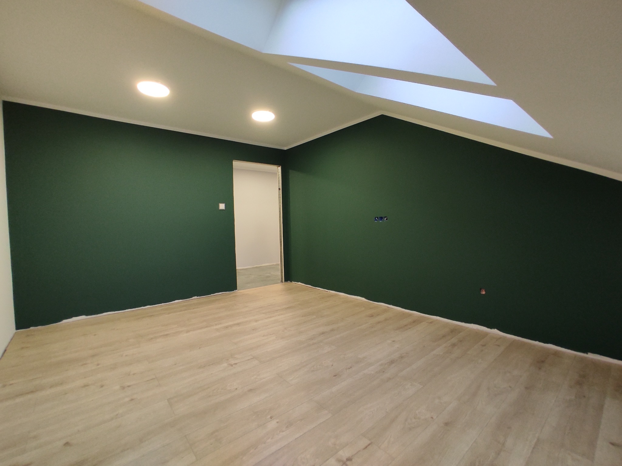 Poddasze po remoncie: ściany pomalowane na ciemnozielony kolor, jasne panele podłogowe, widoczne punkty oświetleniowe w suficie i fragment okna dachowego.