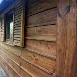 MAREK KĘDZIA BASKAR - Zewnętrzny widok drewnianej altany z oknem z okiennicami, widoczne słoje drewna i konstrukcja z belek.