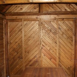 MAREK KĘDZIA BASKAR - Wnętrze drewnianej konstrukcji z ukośnymi deskami na ścianie, widoczne elementy dachu i stolarka okienna z widokiem na zieleń.