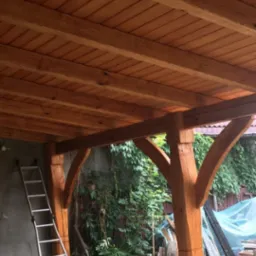 Konstrukcja z belek i drewnianego zadaszenia, widoczne mocne słupy podtrzymujące, drabina oparta o ścianę, roślinność w tle, surowy stan wykończenia posadzki.