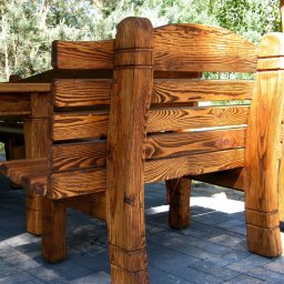 MAREK KĘDZIA BASKAR - Rustykalny zestaw mebli ogrodowych z drewna o wyraźnym usłojeniu, składający się ze stołu i ław, umieszczony na brukowanej nawierzchni w otoczeniu zieleni.