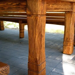 MAREK KĘDZIA BASKAR - Fragment konstrukcji drewnianej altany lub pergoli z widocznymi słupami nośnymi o wyraźnej strukturze drewna, ustawionej na szarym brukowanym podłożu.