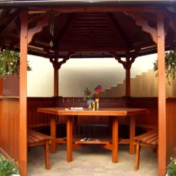 Drewniana altana z ciemnobrązowym dachem i ściankami, wyposażona w stół z ławkami, ozdobiona wiszącymi kwiatami w doniczkach i wiatraczkiem na stole.