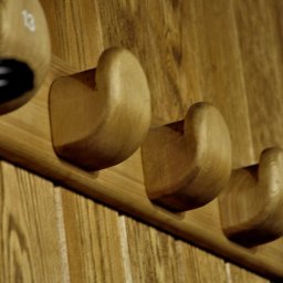 MAREK KĘDZIA BASKAR - Drewniany wieszak ścienny z czterema uchwytami w kształcie zaokrąglonych kostek, jeden z uchwytów z czarnym elementem, całość zamontowana na panelu z jasnego drewna.