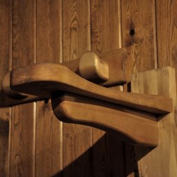 MAREK KĘDZIA BASKAR - Fragment drewnianej konstrukcji z zaokrąglonymi elementami, przymocowanej do boazerii w ciepłym oświetleniu.