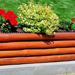 Rustykalna donica z drewnianych bali, wypełniona ziemią i obsadzona czerwonymi pelargoniami oraz krzewami o żółto-zielonych liściach, ustawiona na betonowym obrzeżu trawnika.