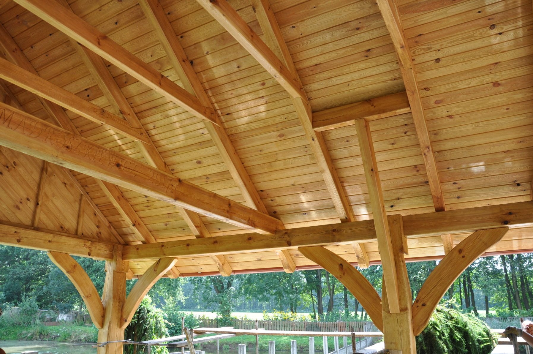 Wnętrze drewnianej altany z widoczną konstrukcją dachu, jasne drewno, inskrypcje na belkach, widok na zewnątrz na zieleń.