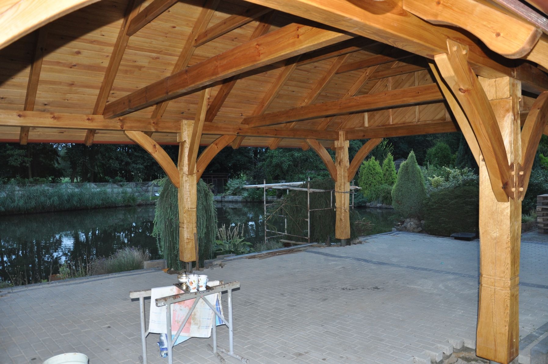 Drewniana konstrukcja zadaszenia z widocznymi elementami łączeń, ustawiona na utwardzonym terenie z widokiem na staw i zieleń w tle; na pierwszym planie prowizoryczny stolik z naczyniami.