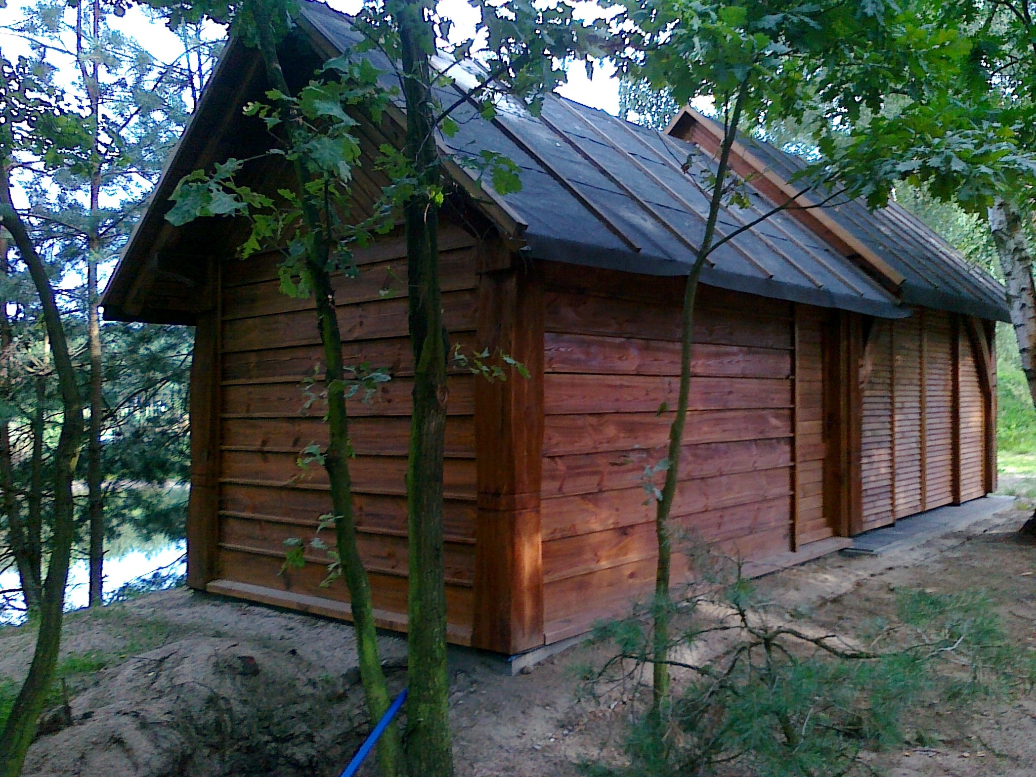 Rustykalna, drewniana konstrukcja z ciemnym dachem, otoczona drzewami, z widokiem na jezioro w tle.