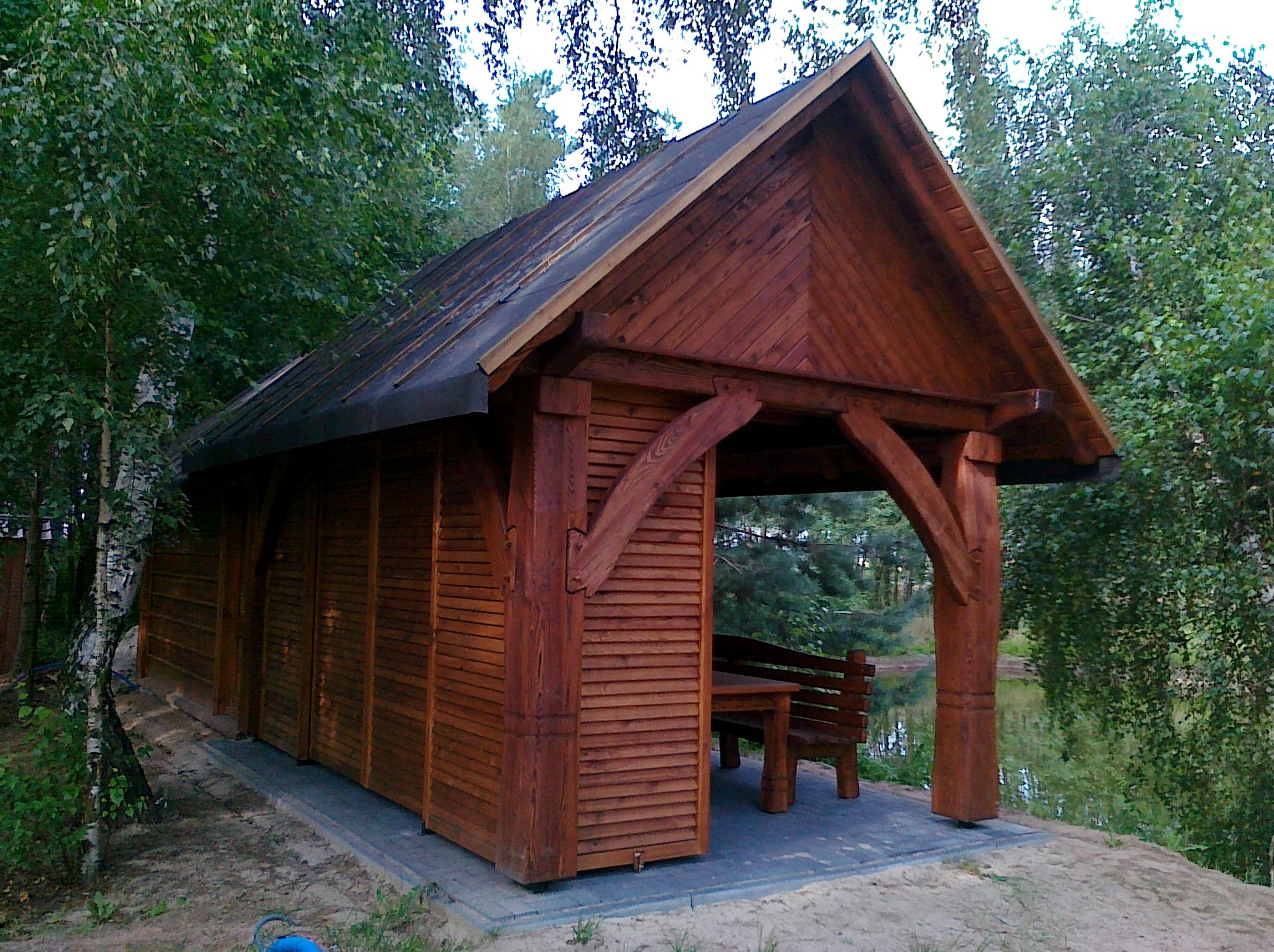 Zadaszona konstrukcja drewniana ze stołem i ławką, prawdopodobnie w Słupii Kapitulnej, z widokiem na staw w tle, otoczona drzewami.