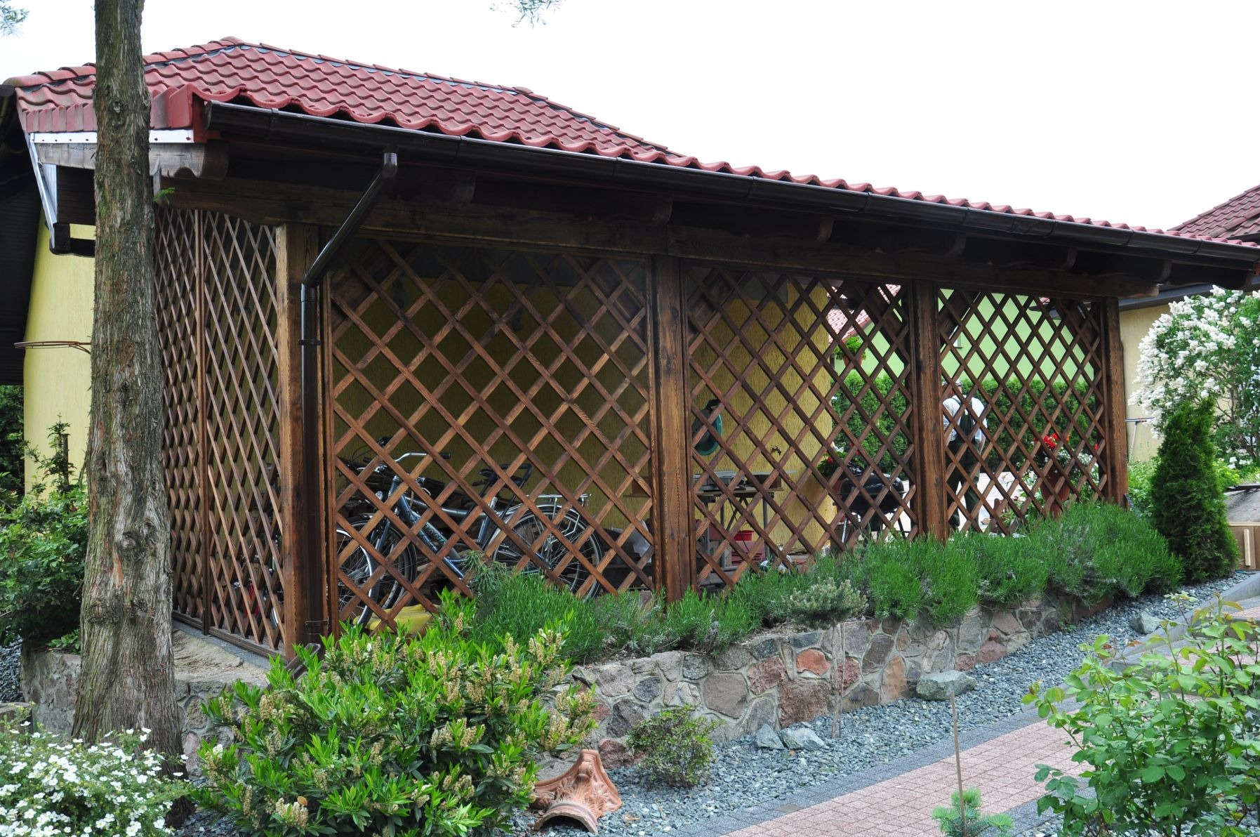 Zadaszona konstrukcja z drewnianą kratą, prawdopodobnie wiata rowerowa, obok drzewo i kamienny murek z zielenią
