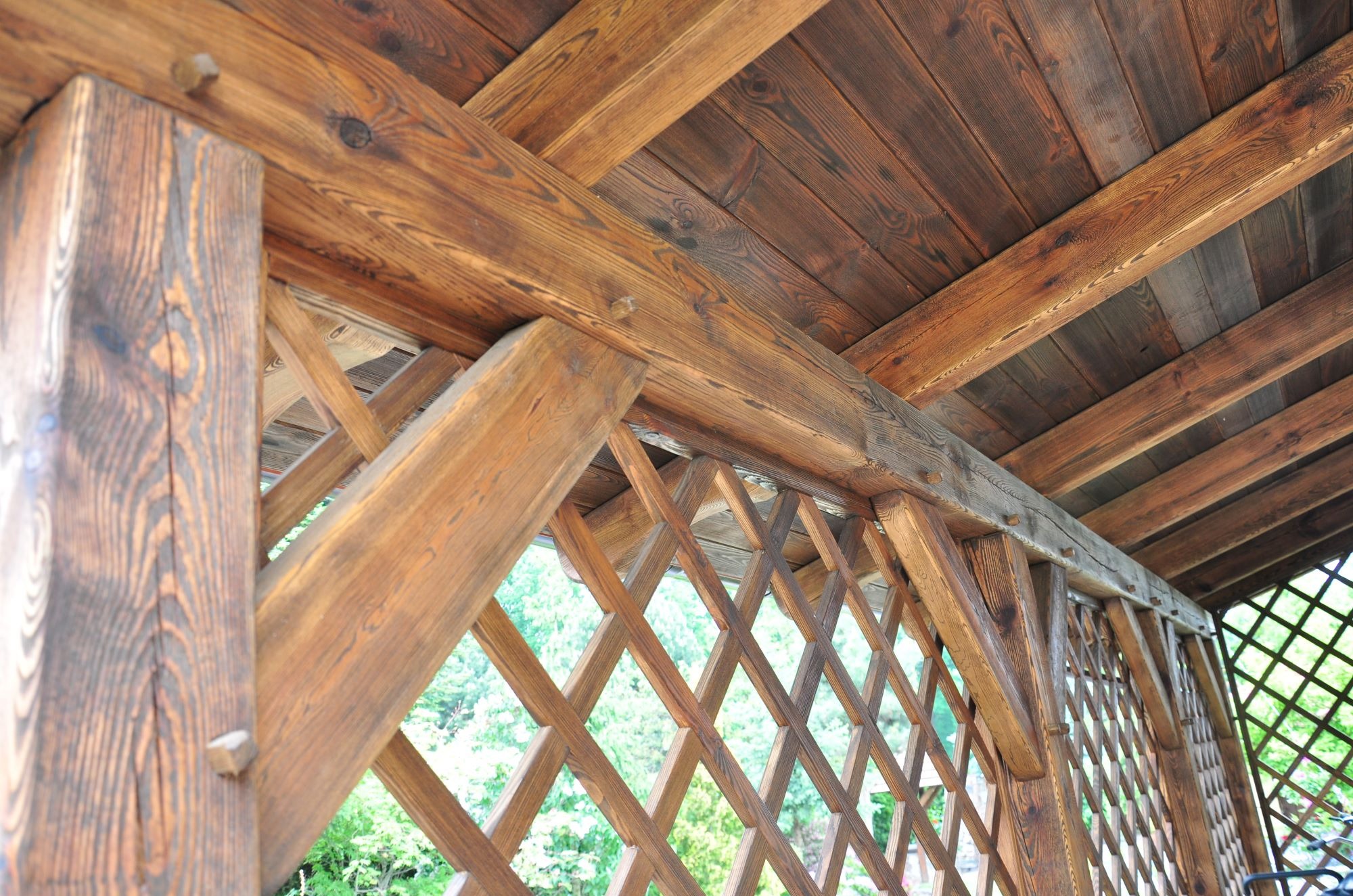 Fragment drewnianej konstrukcji z belek i kratownicy, widok od dołu, ukazujący połączenia elementów za pomocą kołków, z widokiem na zieleń w tle.