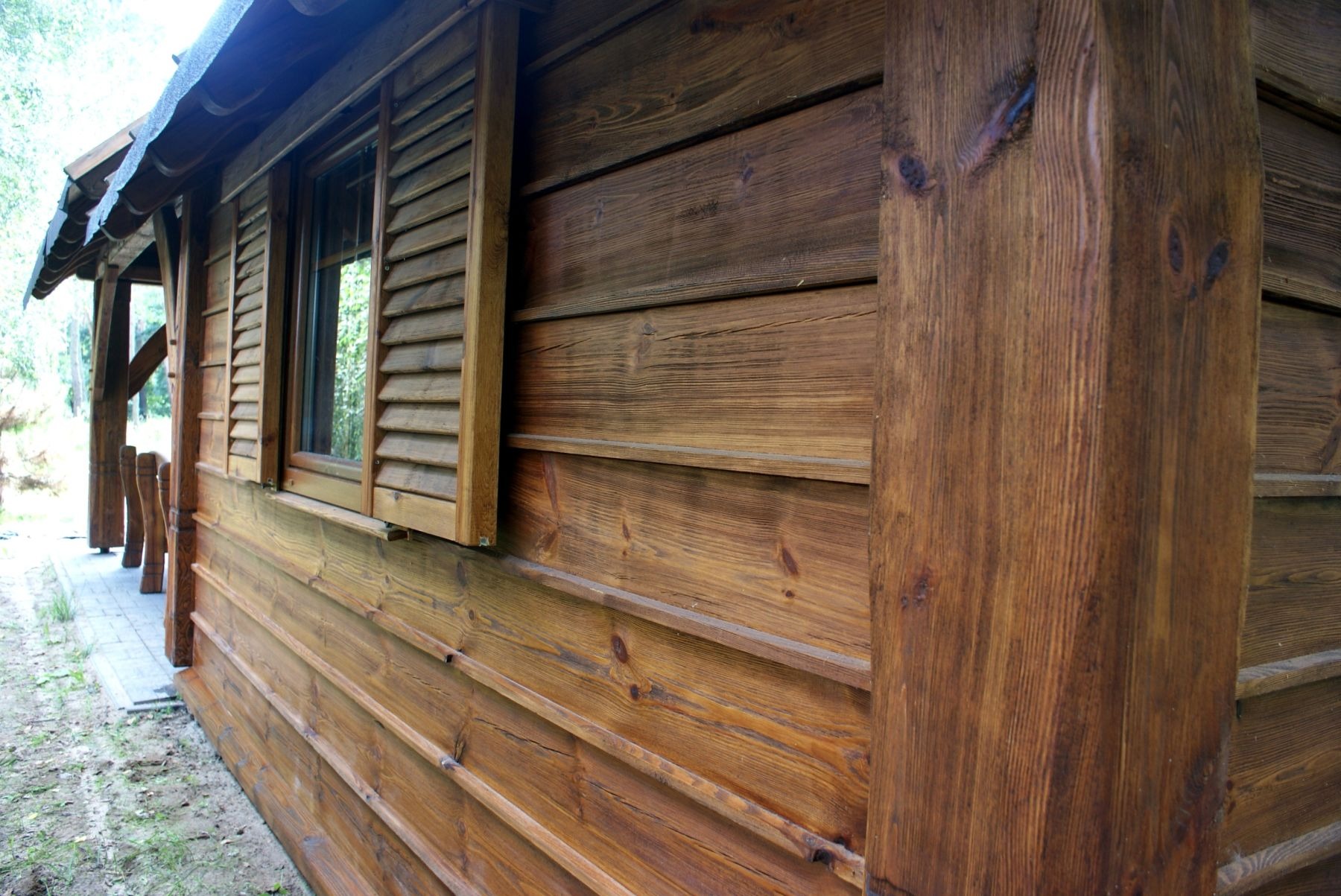 Zewnętrzny widok drewnianej altany z oknem z okiennicami, widoczne słoje drewna i konstrukcja z belek.