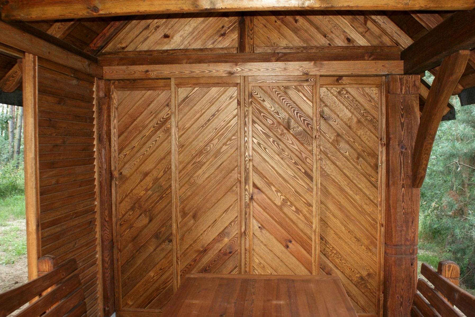 Wnętrze drewnianej konstrukcji z ukośnymi deskami na ścianie, widoczne elementy dachu i stolarka okienna z widokiem na zieleń.