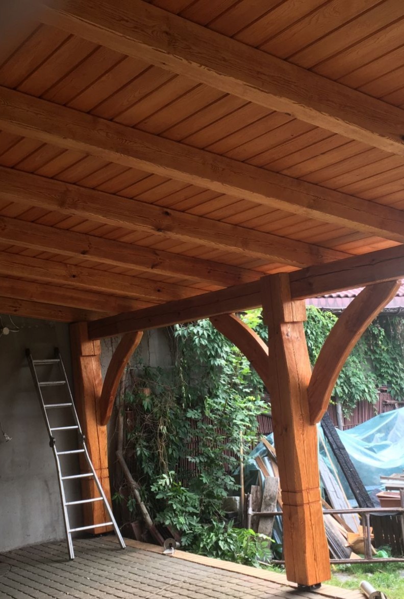 Konstrukcja z belek i drewnianego zadaszenia, widoczne mocne słupy podtrzymujące, drabina oparta o ścianę, roślinność w tle, surowy stan wykończenia posadzki.