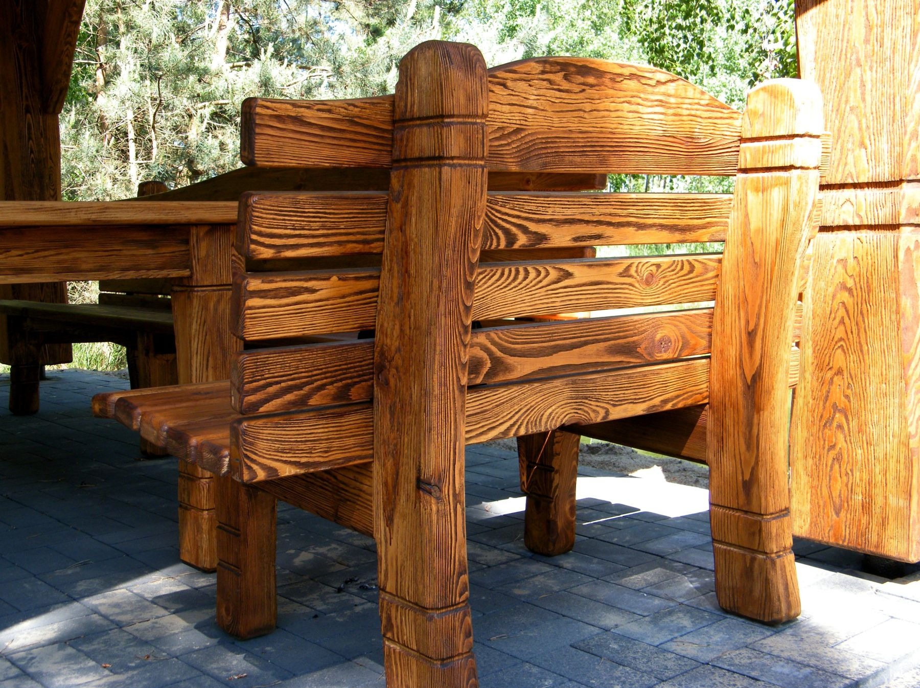 Rustykalny zestaw mebli ogrodowych z drewna o wyraźnym usłojeniu, składający się ze stołu i ław, umieszczony na brukowanej nawierzchni w otoczeniu zieleni.