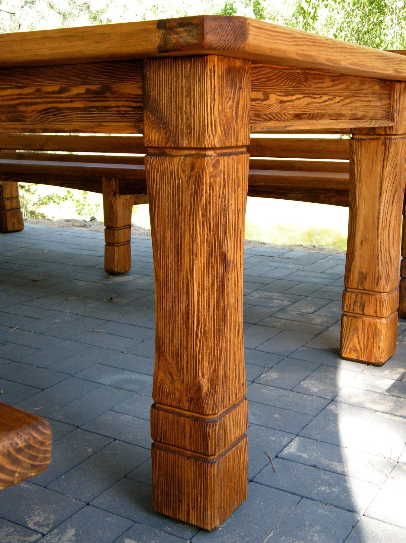 Fragment konstrukcji drewnianej altany lub pergoli z widocznymi słupami nośnymi o wyraźnej strukturze drewna, ustawionej na szarym brukowanym podłożu.