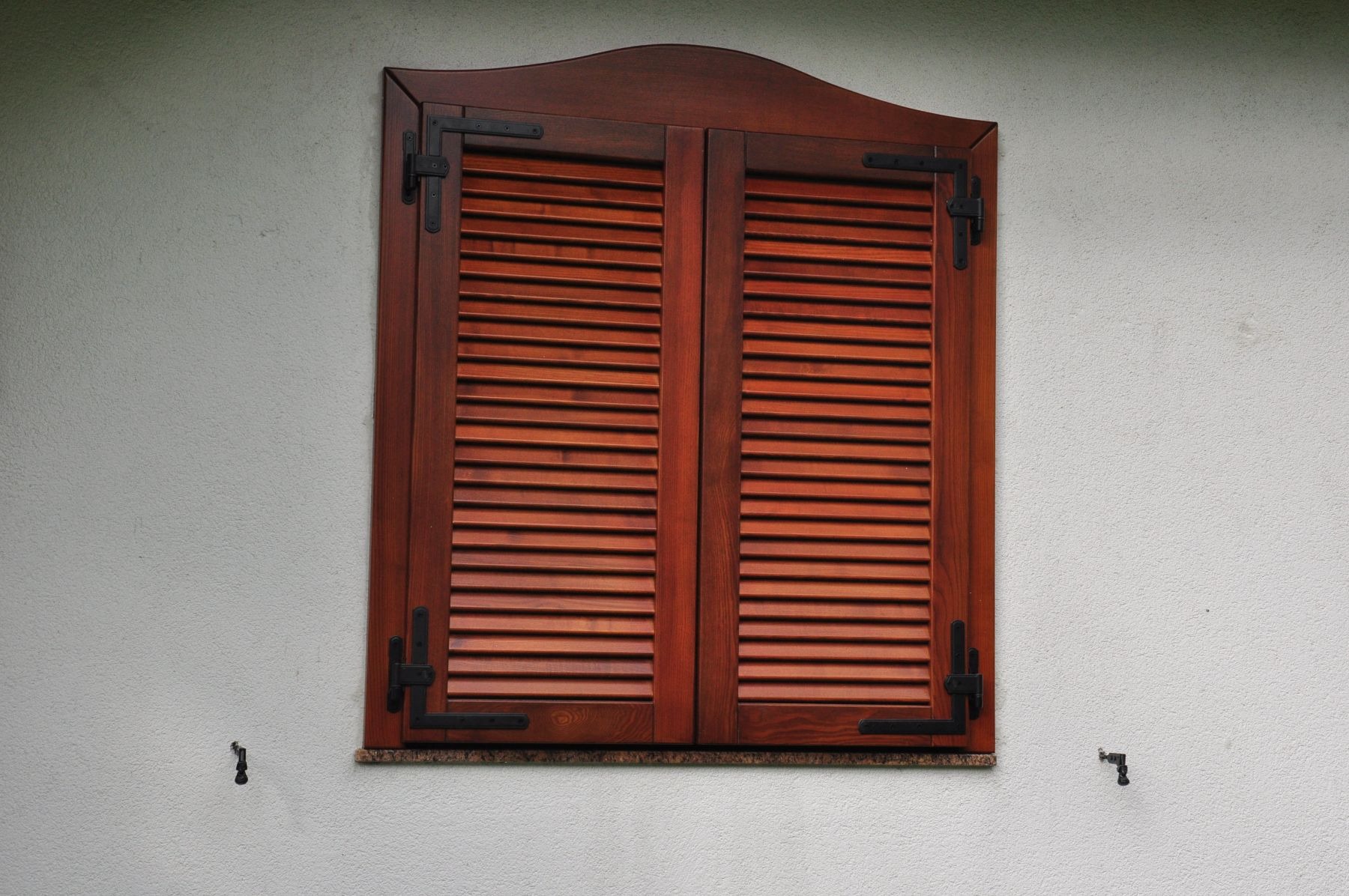 Zewnętrzne okiennice drewniane w kolorze ciepłego brązu z czarnymi zawiasami na białej, teksturowanej ścianie.