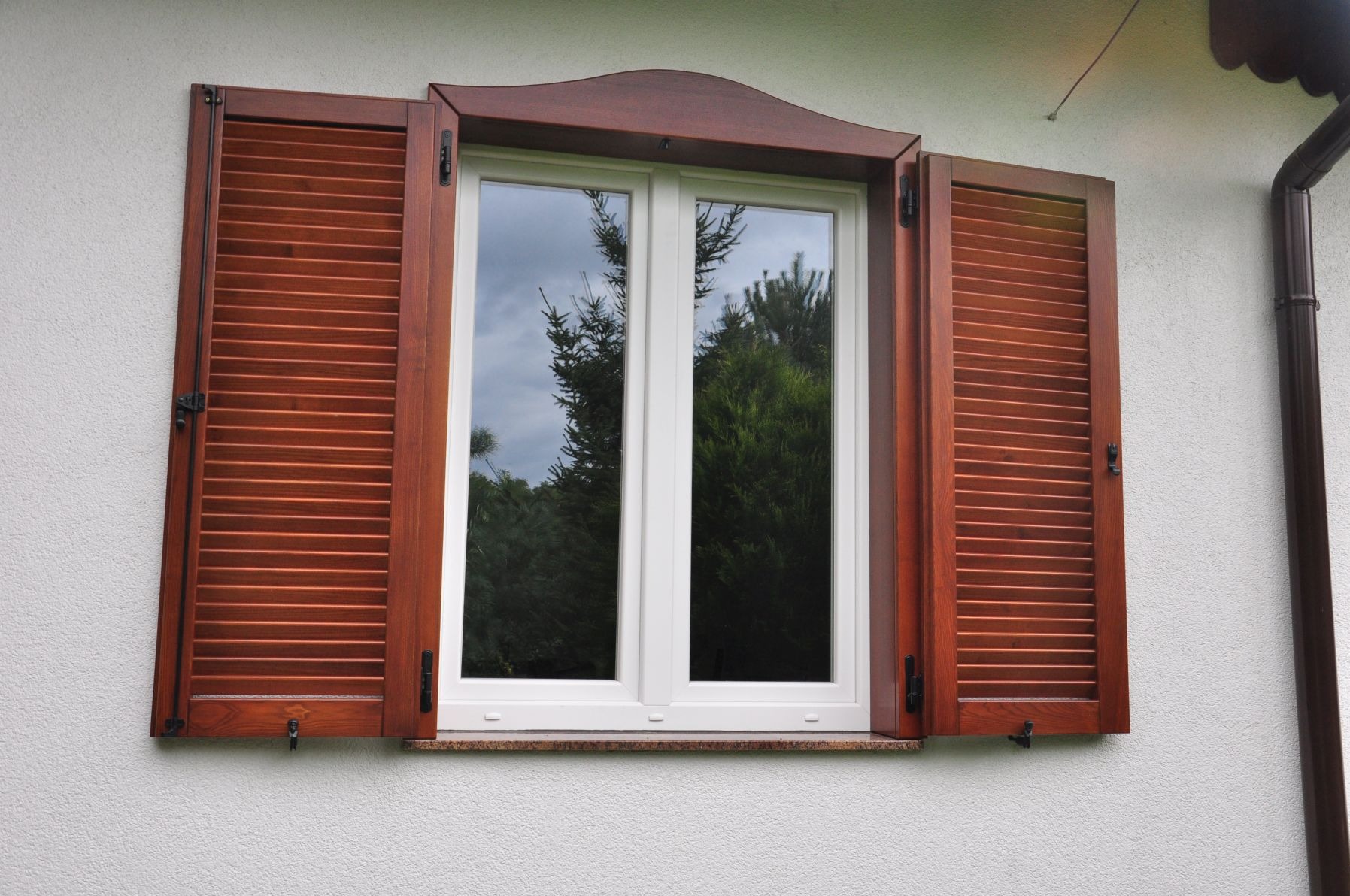 Okna z drewnianymi okiennicami żaluzjowymi w kolorze orzechowym, zamontowane na białej elewacji domu z widocznym fragmentem rynny.