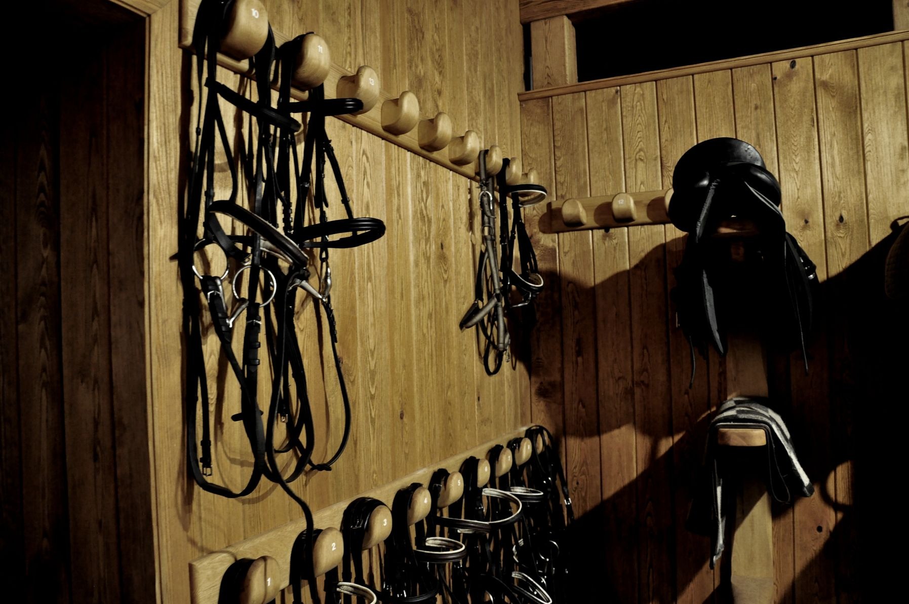 Uprzęże i siodła jeździeckie zawieszone na drewnianych wieszakach przytwierdzonych do boazerii w stajni.