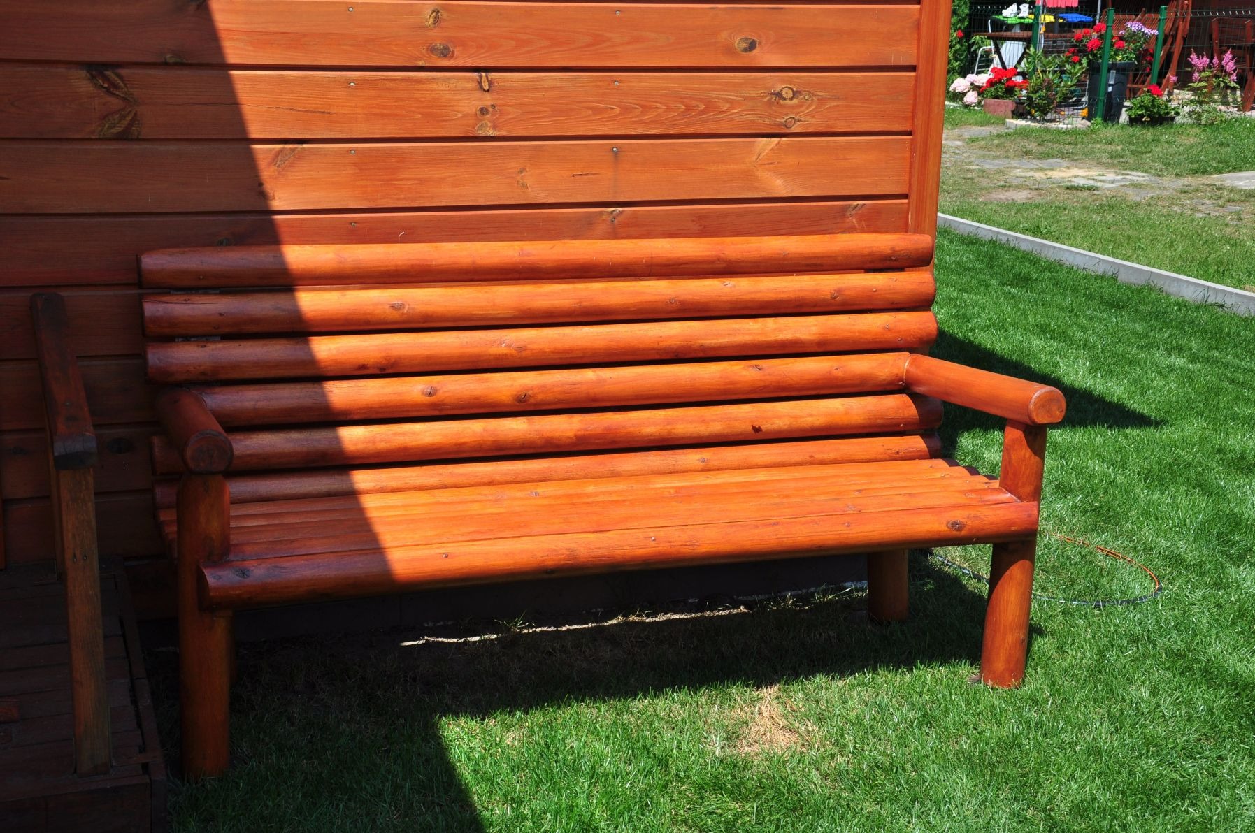 Drewniana ławka ogrodowa z bali, pomalowana na brązowo, ustawiona na trawniku przed ścianą z desek, częściowo zacieniona.