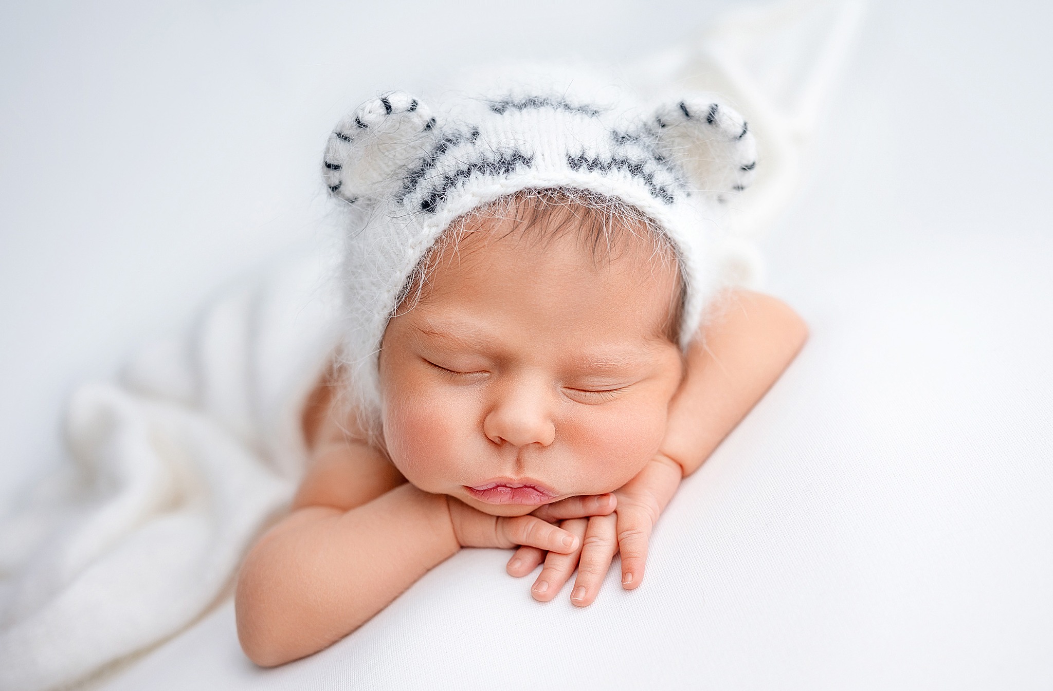 Śpiący noworodek w białym, puszystym stroju z uszkami, leżący na białym tle, z rączkami pod policzkiem.