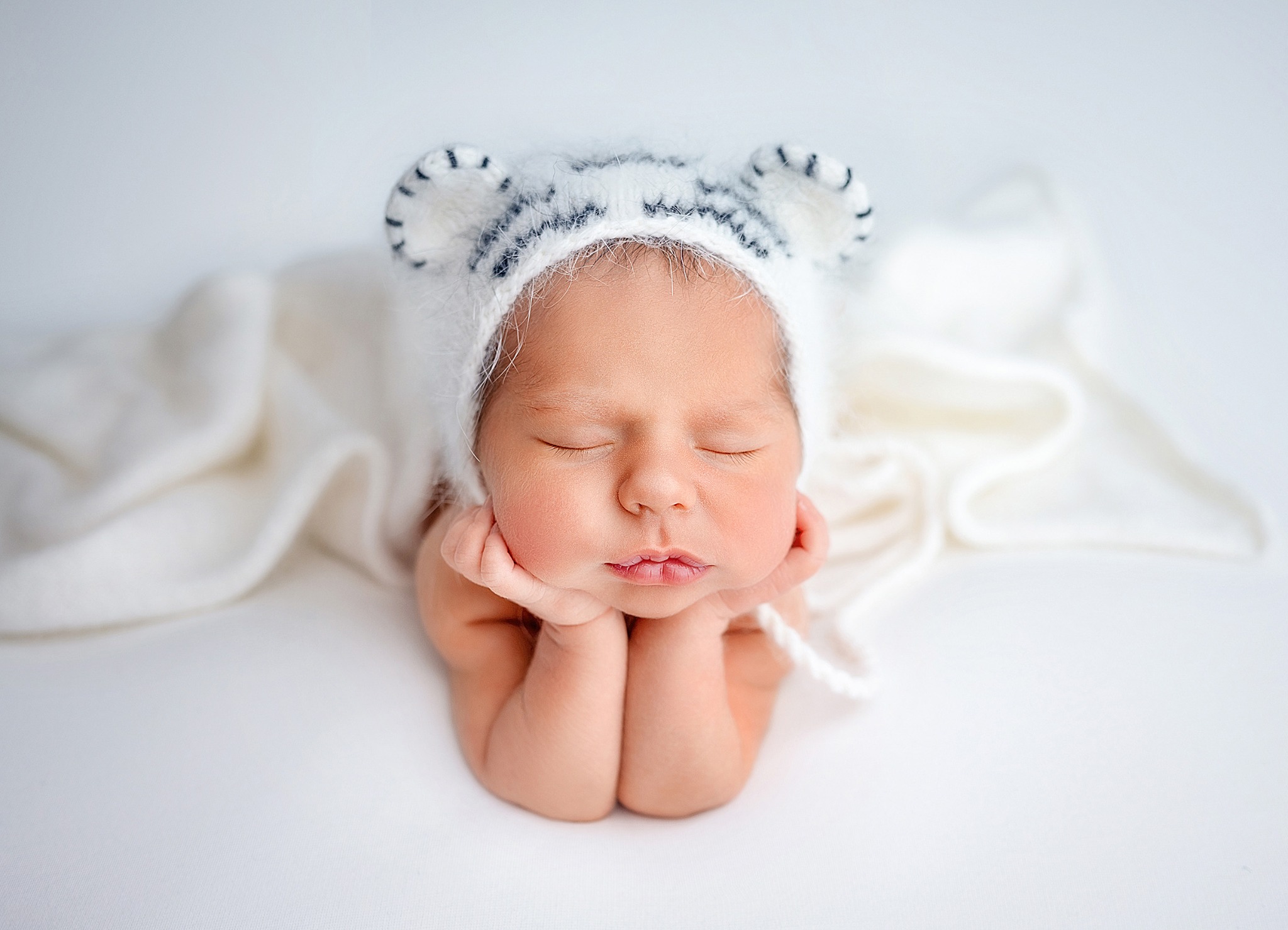 Śpiący noworodek w białym, puszystym czepku z uszkami w paski, delikatnie podpierający główkę rączkami, na białym tle.