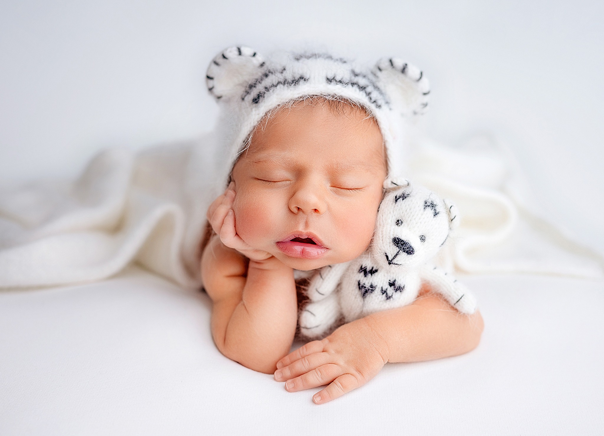 Śpiący noworodek w białym, dzierganym stroju tygryska, tulący pluszowego tygryska na białym tle.