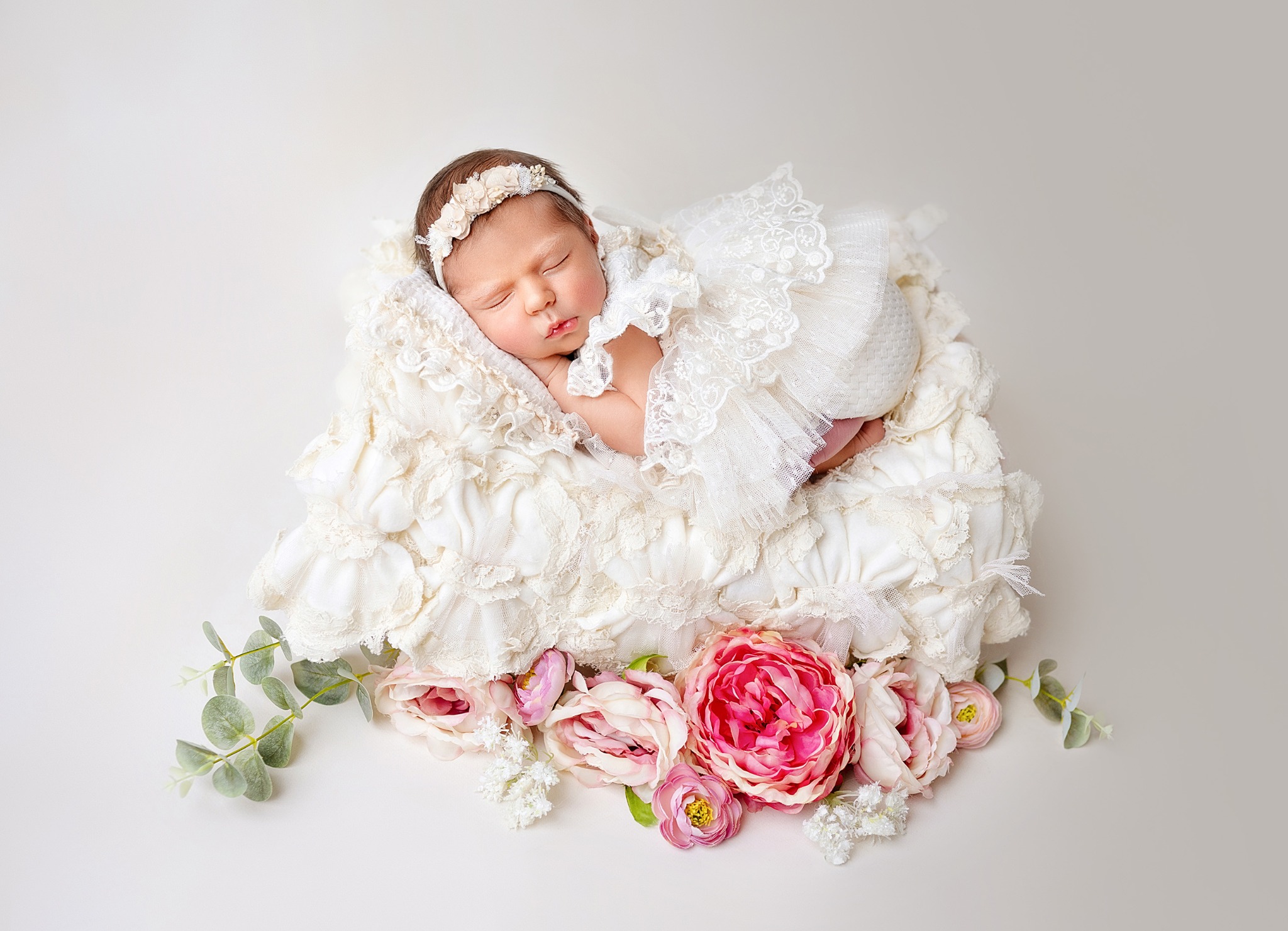 Noworodek w koronkowej sukience i opasce na głowie śpiący na puszystym, kremowym kocyku z dekoracją z różowych i białych kwiatów na jasnym tle.