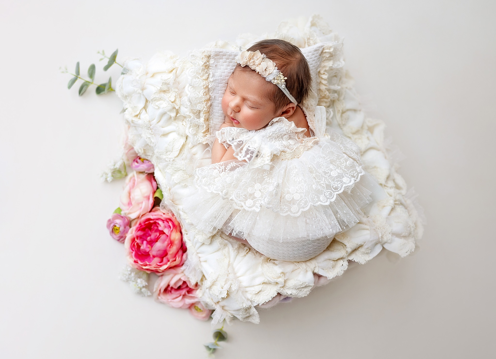 Śpiąca noworodka w białej koronkowej sukience i opasce z kwiatami, ułożona na ozdobnej poduszce z kwiatami i zielonymi gałązkami na jasnym tle.