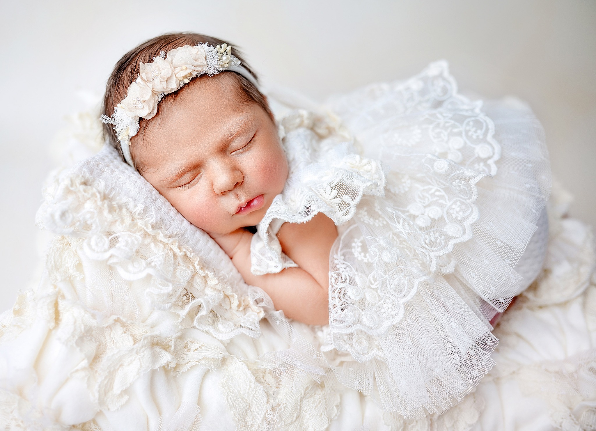 Śpiąca noworodka w koronkowej sukience i opasce z kwiatami, sfotografowana w Krakowie na miękkiej poduszce z falbankami, w jasnym, naturalnym oświetleniu.