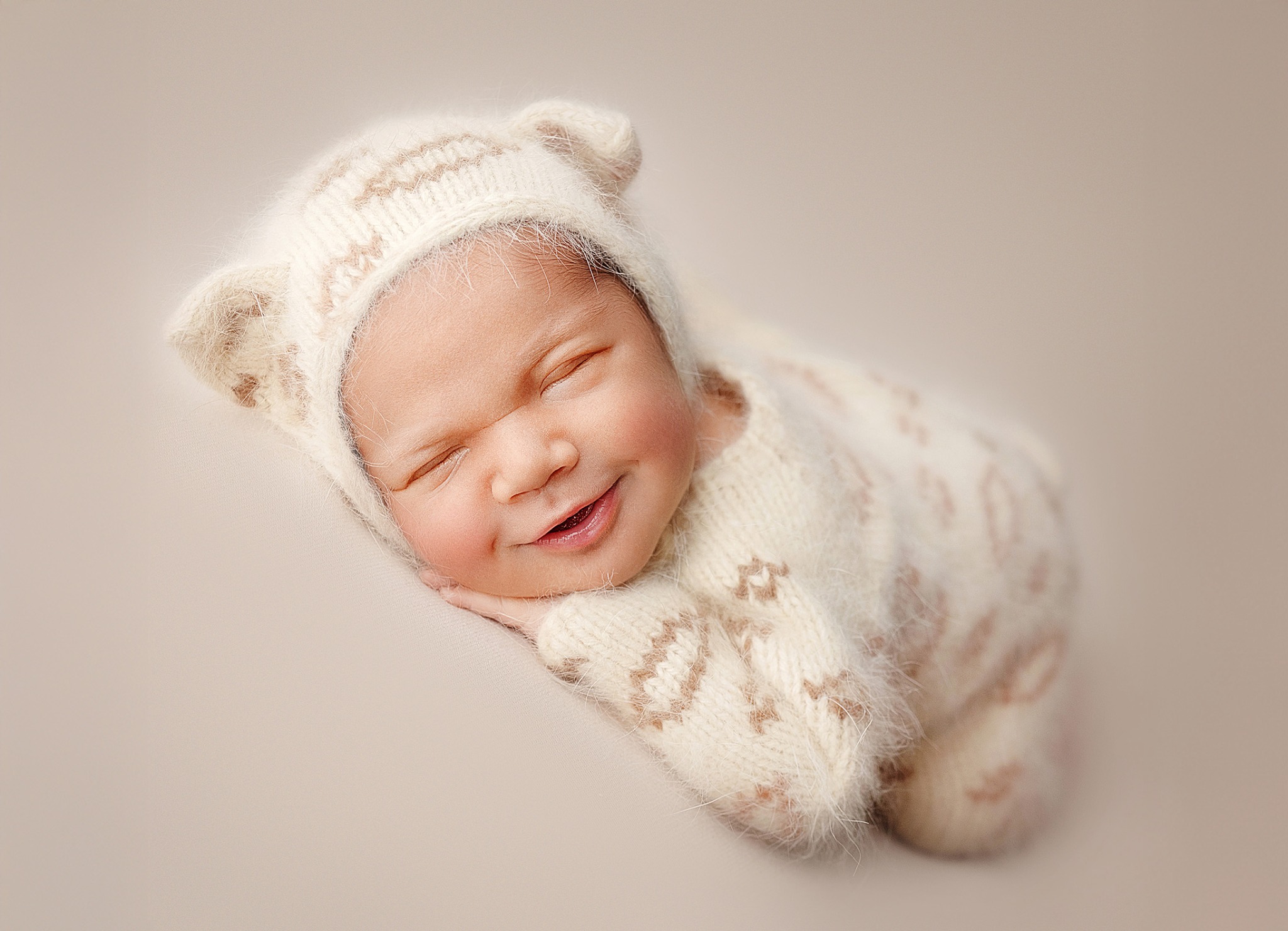 Śpiący noworodek w kremowym, dzierganym kombinezonie z uszkami na beżowym tle, delikatny uśmiech na twarzy.