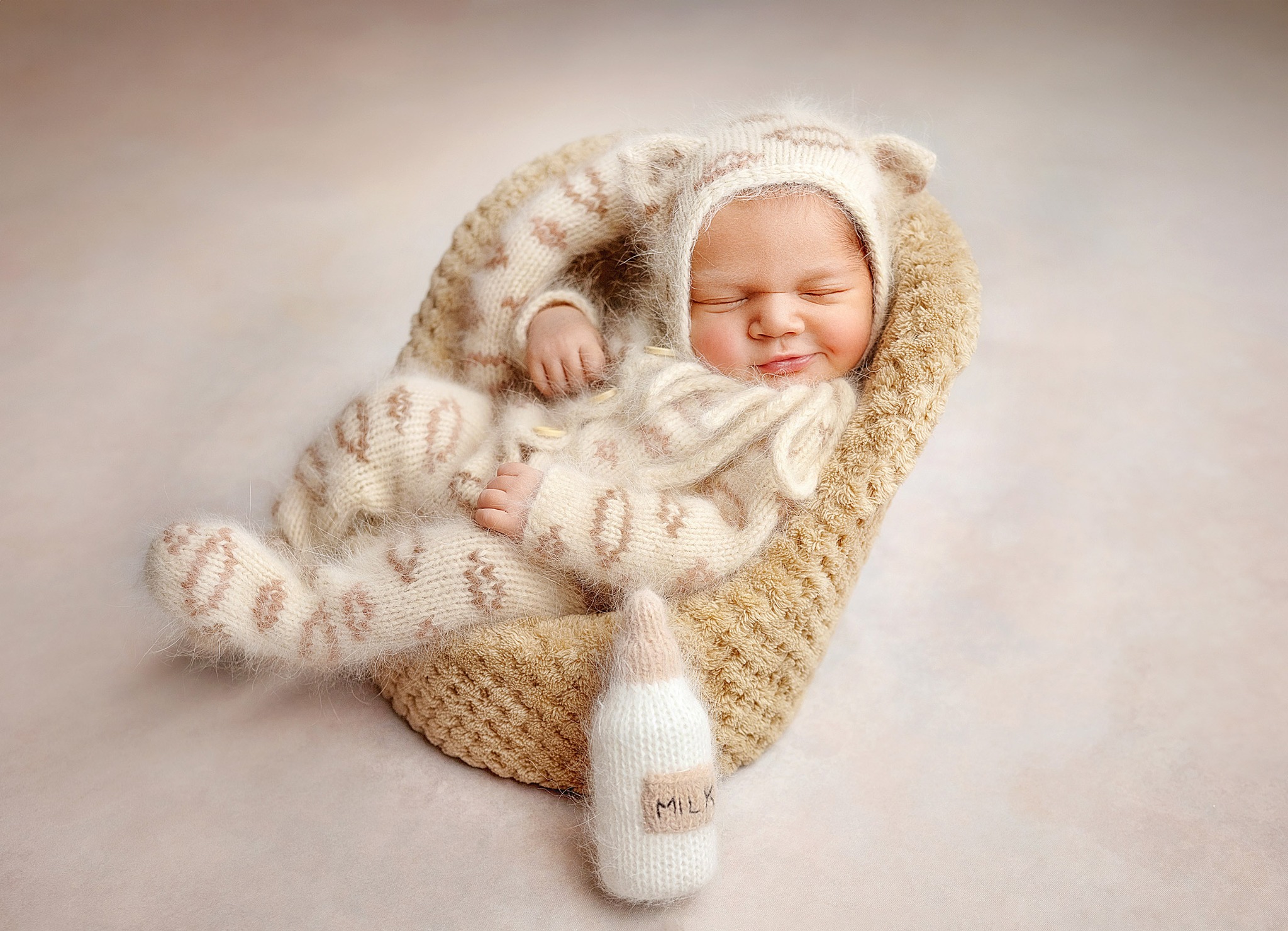 Uśmiechnięty noworodek śpiący w beżowym, dzierganym kombinezonie z uszkami i w dzierganym gniazdku, obok stoi dziergana butelka z napisem MILK.