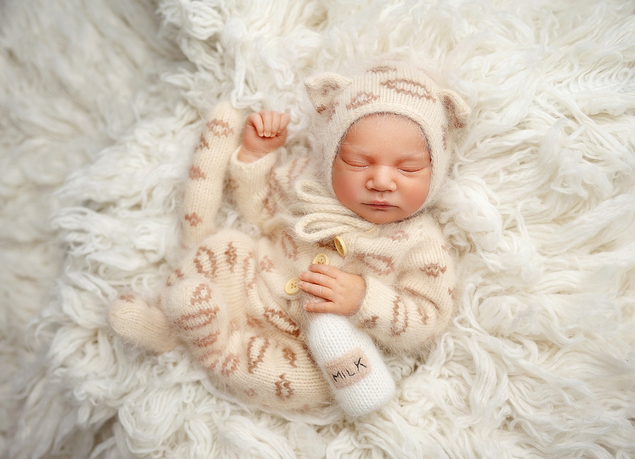 Śpiący noworodek w beżowym, dzierganym kombinezonie z kapturem z uszkami, leżący na puszystym, białym tle, trzymający w dłoni dzierganą butelkę z napisem MILK.