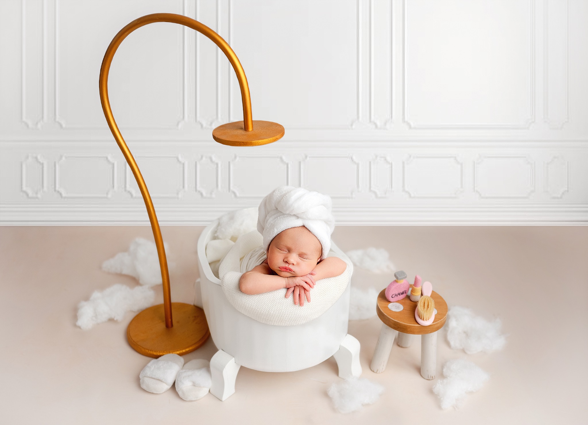 Noworodek w białym turbanie śpiący w małej białej wanience z rekwizytami do kąpieli, oświetlony złotą lampą, na tle białej ściany z panelami i beżowej podłogi z dekoracyjnymi chmurkami.