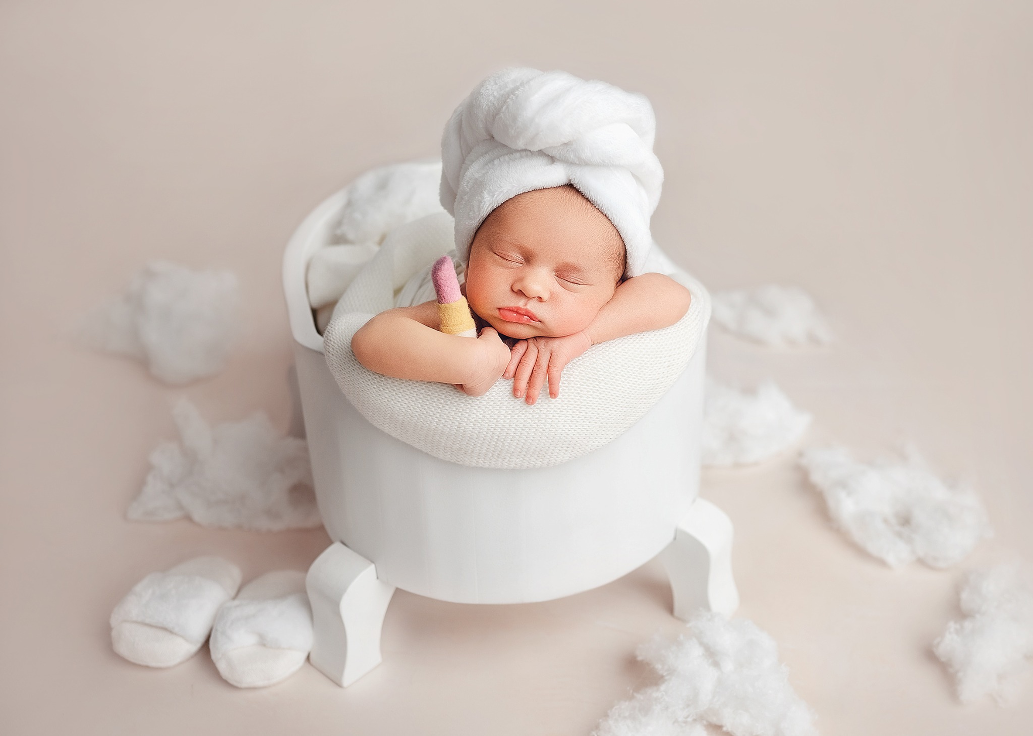 Noworodek śpiący w białej wanience z ręcznikiem na głowie, trzymający filcową zabawkę w kształcie szminki, otoczony białymi obłoczkami i pluszowymi kapciami.