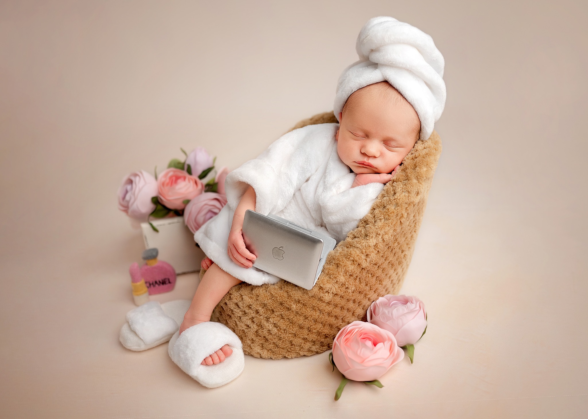 Noworodek w białym szlafroku i turbanie na głowie, śpiący w pluszowym fotelu, trzymając srebrnego laptopa Apple, obok kwiaty i pantofle Chanel.