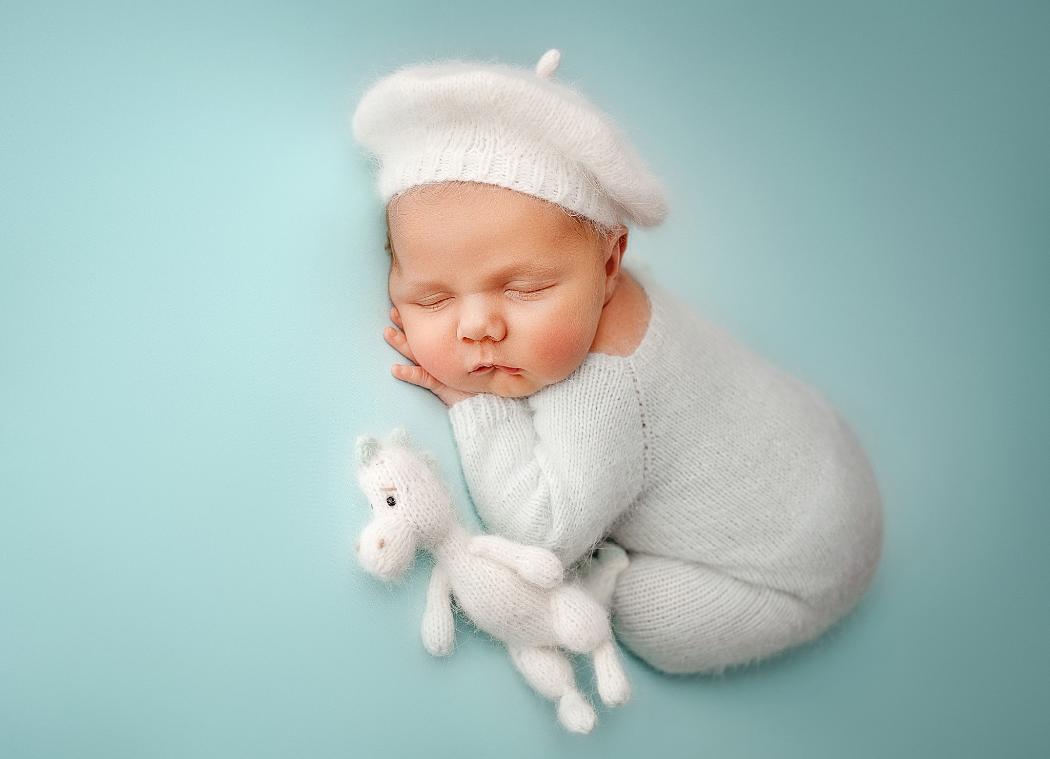Śpiący noworodek w białym, dzierganym berecie i kombinezonie, leżący na boku z dzierganym konikiem na błękitnym tle.