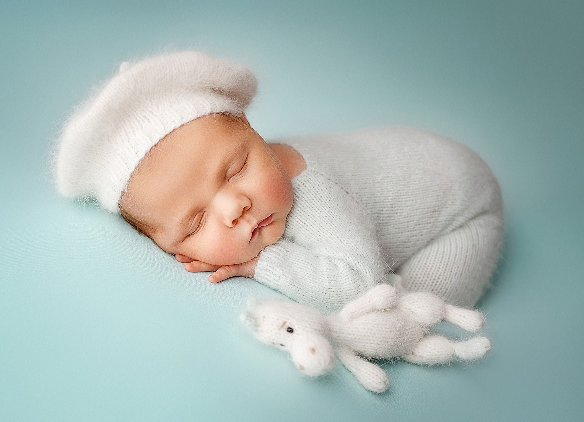 Śpiący noworodek w białym beretce i kombinezonie, leżący na boku obok pluszowego konika na turkusowym tle, delikatne oświetlenie.