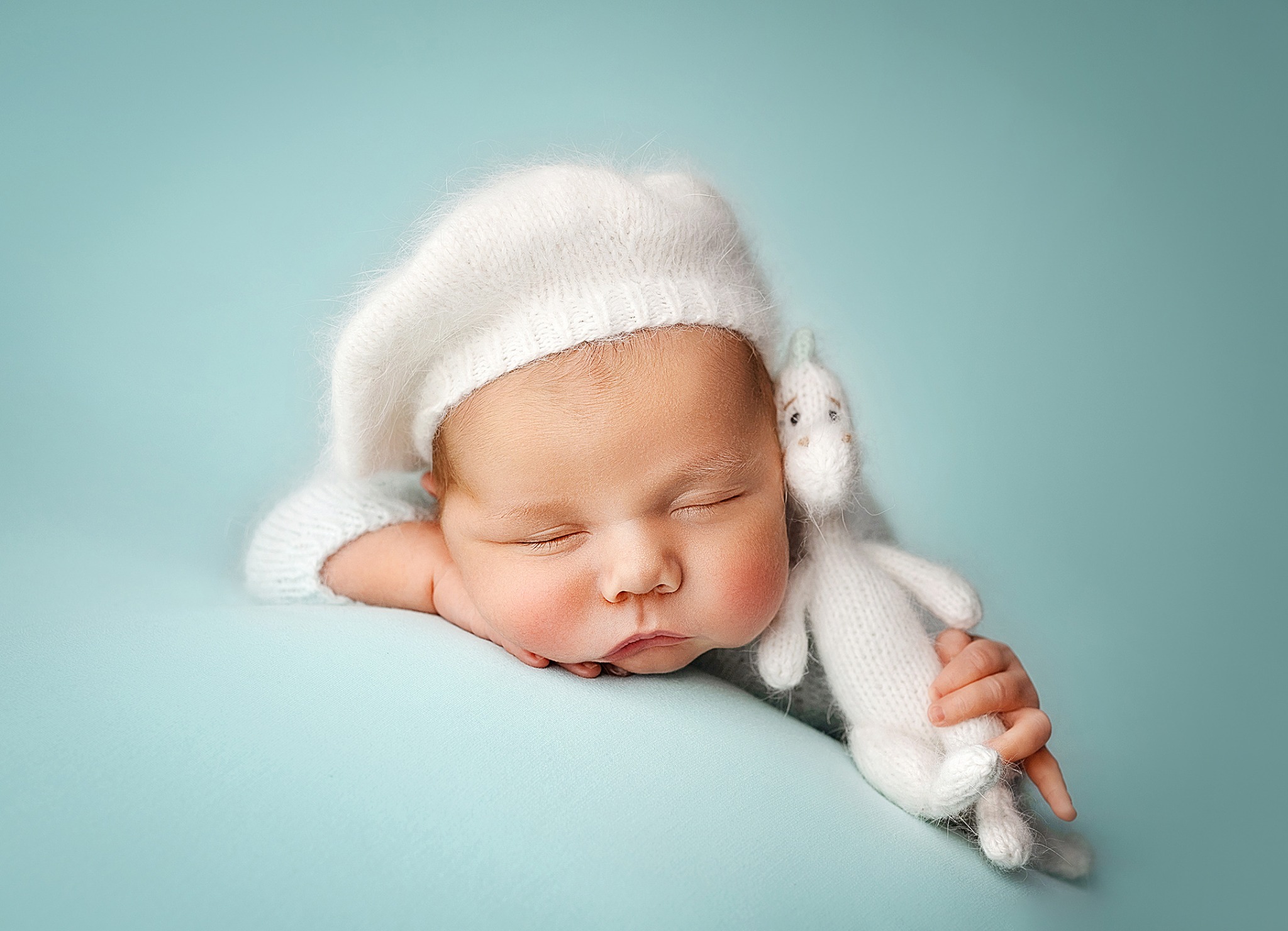 Śpiący noworodek w białej czapce, przytulający dzierganą zabawkę królika na jasnoniebieskim tle.