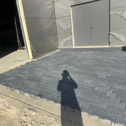 STATUS-BRUK Petro Tsymbaliak - Fragment nowej, szarej nawierzchni z kostki brukowej ułożonej przed budynkiem przemysłowym z szarymi drzwiami, w tle widoczne materiały budowlane i cień osoby robiącej zdjęcie.