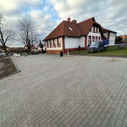 STATUS-BRUK Petro Tsymbaliak - Nowa, szara nawierzchnia z kostki brukowej na tle budynku szkoły z czerwoną dachówką i zaparkowanym niebieskim samochodem dostawczym.