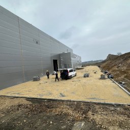 STATUS-BRUK Petro Tsymbaliak - Przygotowanie podłoża piaskiem pod kostkę brukową przed budynkiem magazynowym, widoczni pracownicy i samochód dostawczy, obrzeża chodnika wykończone