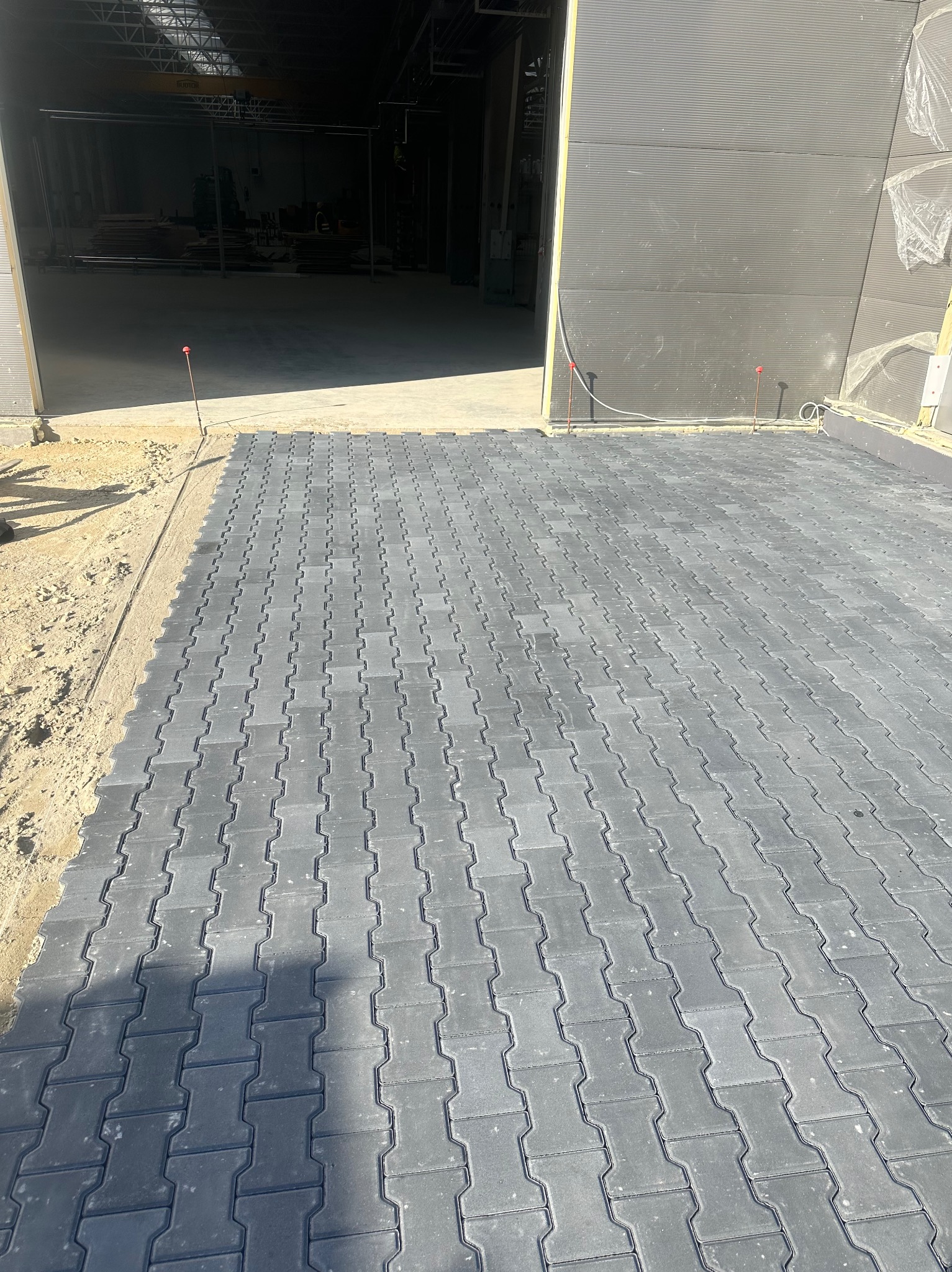 Nowo ułożona, szara kostka brukowa typu 'I', prowadząca do otwartego magazynu z widocznymi elementami konstrukcji stalowej wewnątrz, obramowana piaskiem i zabezpieczona czerwonymi kołkami.